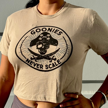 Load image into Gallery viewer, A person wearing a beige cropped T-shirt with a large black pirate skull and crossed swords graphic on the front, with the words 'GOONIES' and 'NEVER SCALE' in the design.
